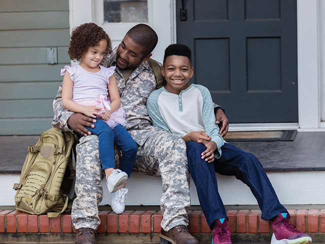man wearing camouflage military fatigues seated on porch with 2 young children 