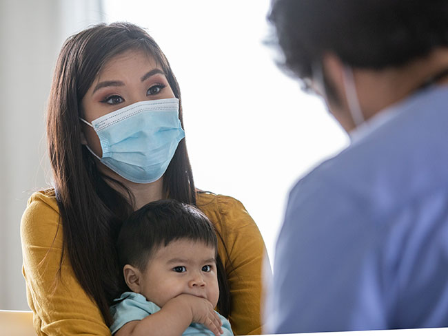 Mother wearing a protective face mask with a baby in her lap, with physician during an exam.