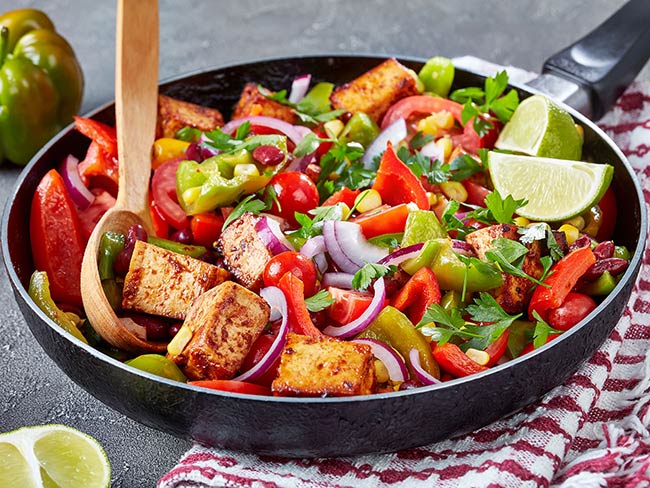 frying pan with tofu and an array of chopped vegetables including bell peppers, red onions, cherry tomatoes, cilantro and corn
