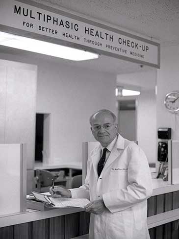 1966 photo of an older male physician standing at a counter in a medical facility.