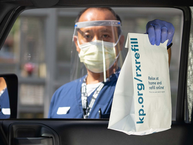 Pharmacy technician wearing face shield handing a bag to a passenger through a car window. 