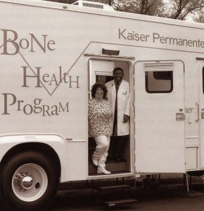 side view of the 'Scan Van' mobile health care vehicle with the door open and Stephen Moki, radiology technologist and health educator, and Pat Brown, clinical assistant standing in the doorway smiling. The words 'Bone Health Program' and 'Kaiser Permanente' are printed on the side.