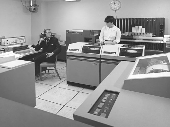 1964 image of a man and woman surrounded by large computers from the time. 
