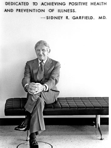 Dr. Garfield seated with legs crossed and smiling in front of a sign that reads "Dedicated to achieving positive health and prevention of illness. Dr. Sidney R Garfield, MD. 