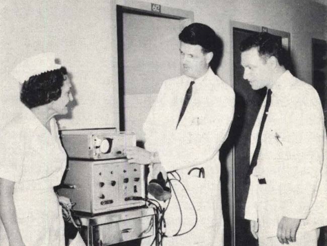 1961 photograph of two male physicians and a nurse standing next to some medical equipment.