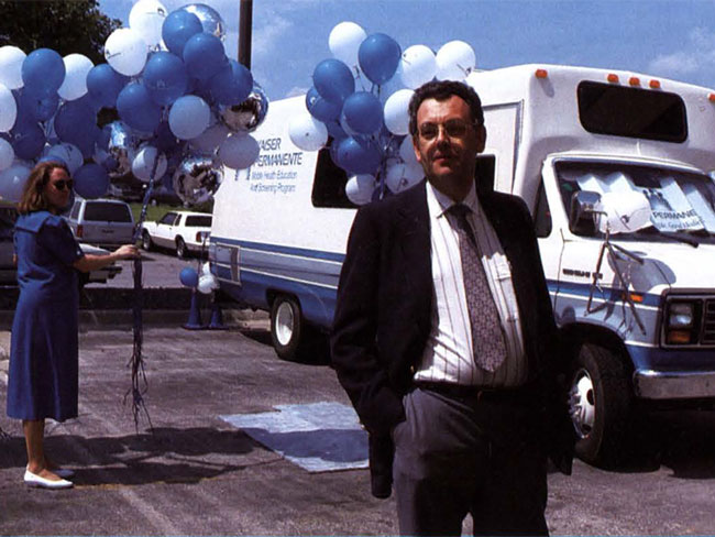man wearing suit and tie standing in front of mobile health care van while woman in the background holds blue and white balloons