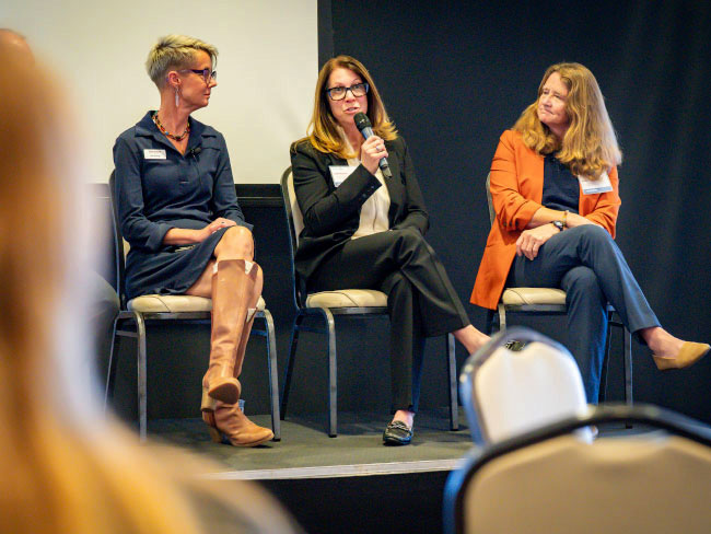 Three women sit on a panel during the Colorado 2025 Community Health Summit, with the woman in the center speaking into a microphone.