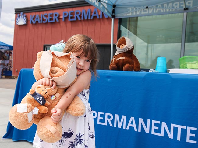 A little girl is hugging a teddy bear in front of a table with a Kaiser Permanente banner