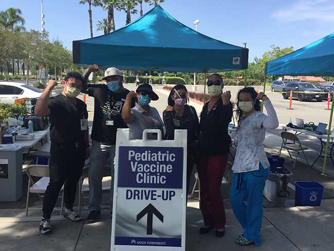 A group of nurses wearing face masks and posing with strong-arms in front of an outdoor pediatric vaccine clinic tent. 