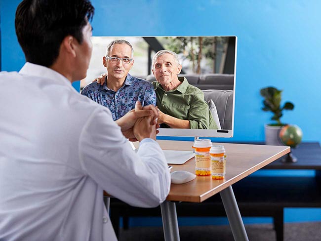 Two men sitting together on a sofa, one with his arm around the other's shoulder, smiling as they have a discussion with a male physician in a video chat.