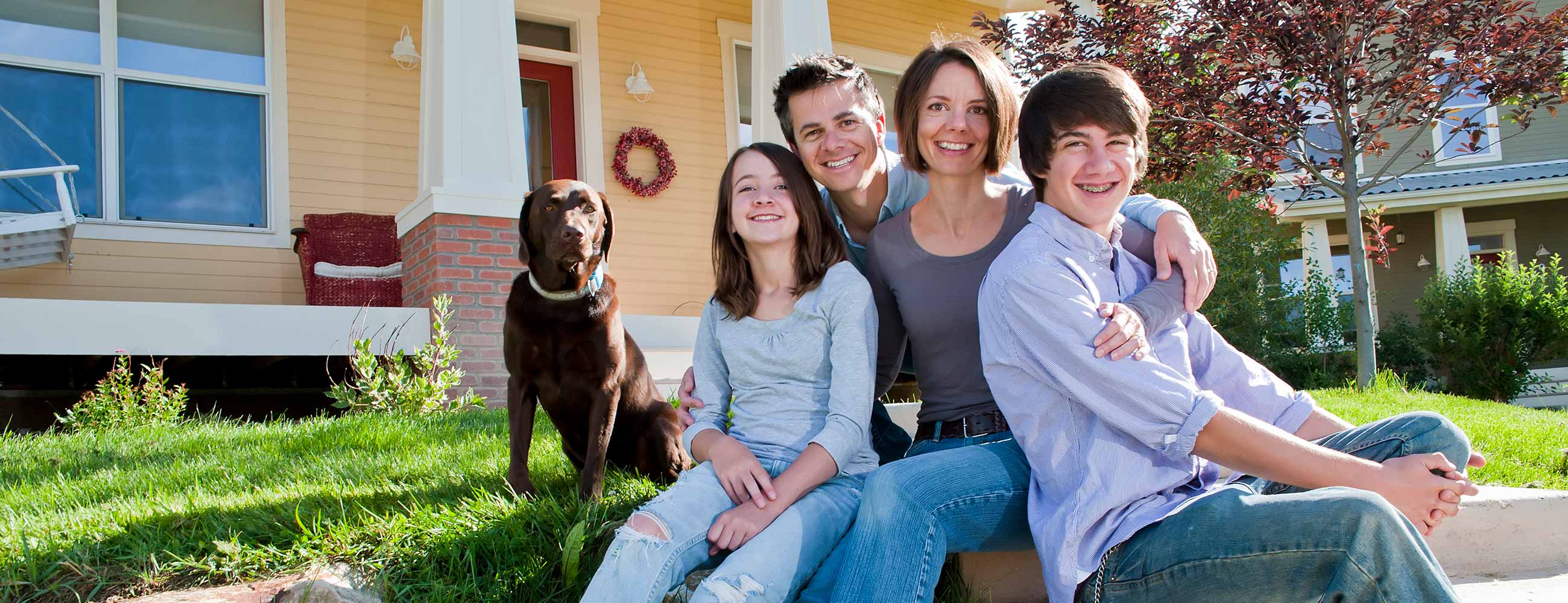  A smiling family, including a mother, father, teenage daughter, and teenage son, sits in the yard in front of their house with their large chocolate lab dog.