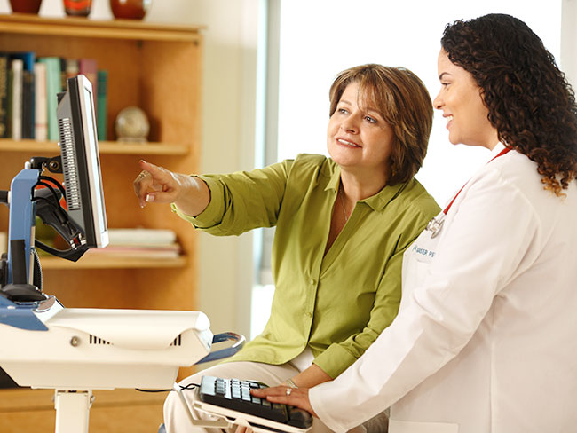 A female physician and her female patient are looking at a computer screen together, with the patient pointing to something on the display.