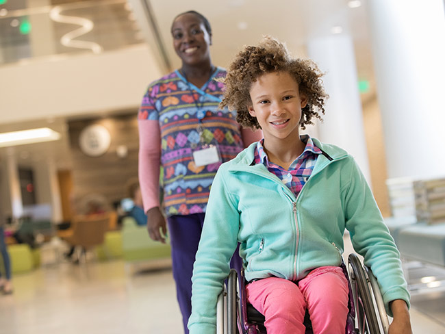 young girl in wheelchair smiling with nurse standing behind her