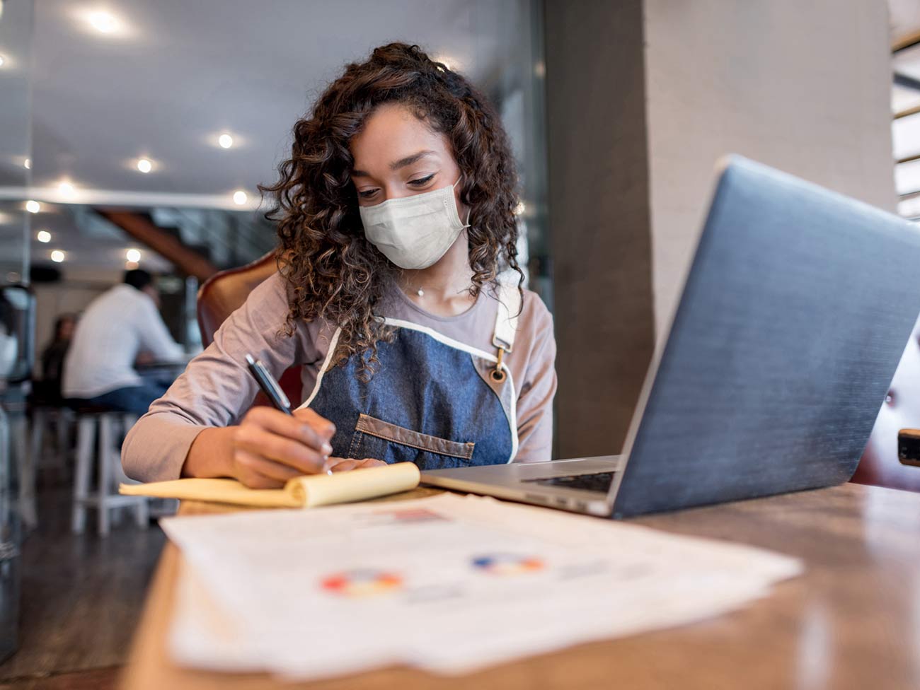 Woman wearing face mask writing in a notepad next to an open laptop.