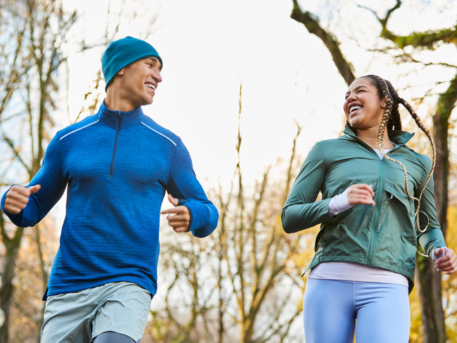 A man and woman smiling at each other as they jog through a wooded area.