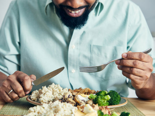 A man eating a healthy protein lunch, made of chicken meat, brown rice, mushrooms, Spanish onion, tomatoes, seeds and broccoli, served in a modern plate on a home wooden table top,