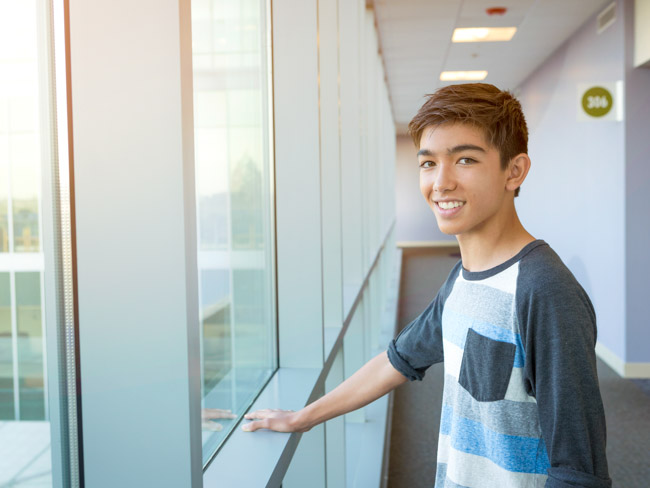 Teenage boy standing by a window in a hallway.