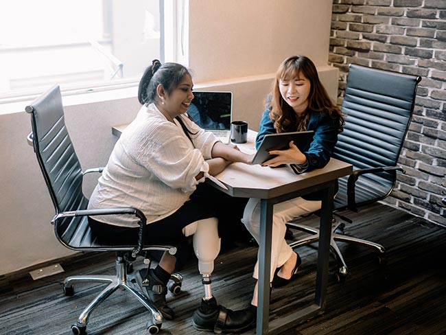 Woman with a prosthetic leg having discussion with her female colleague at an office workstation.