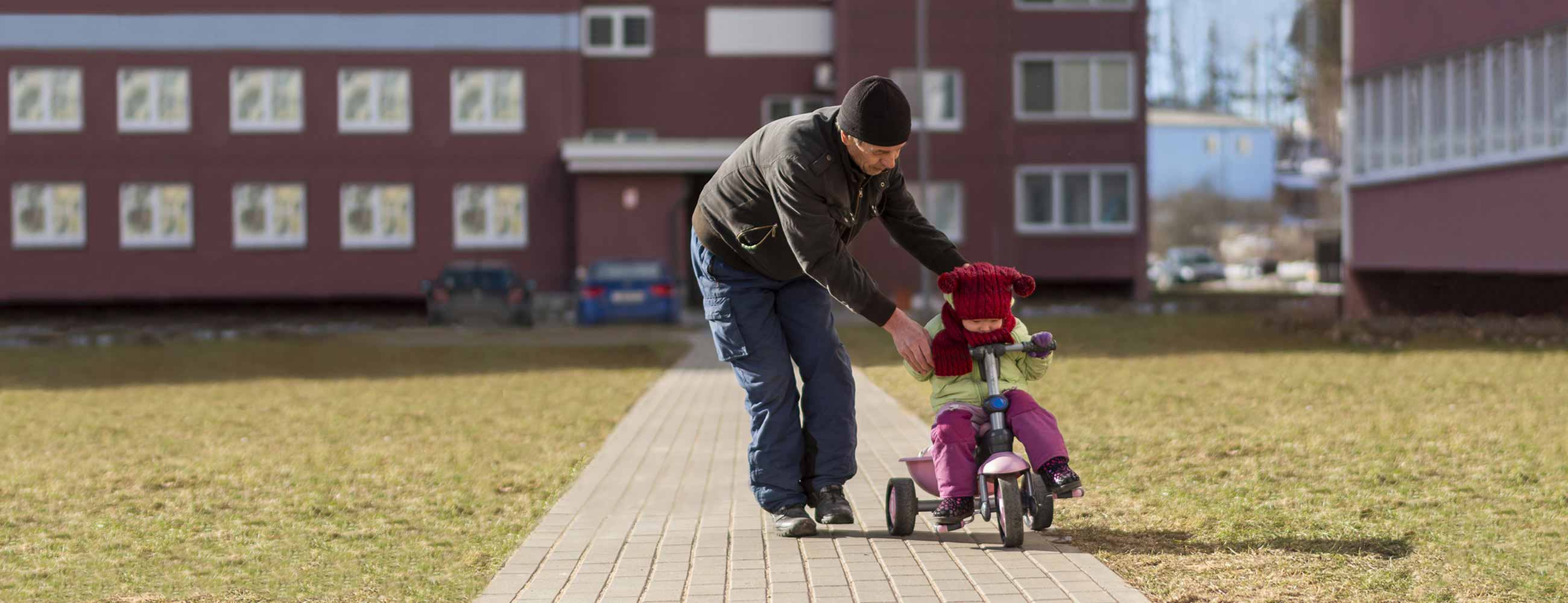 A man teaching his young child how to ride a tricycle out in front of a housing complex. 