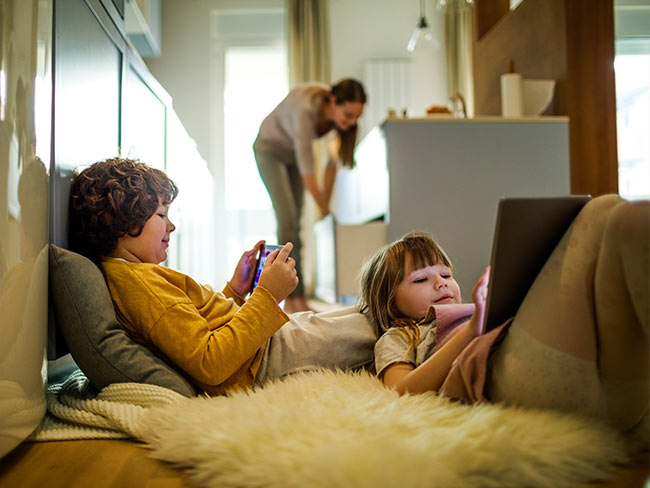 Siblings playing on mobile devices while their mom cleans the kitchen