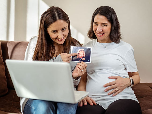 Happy lesbian couple holding ultrasound photo scan on video call.