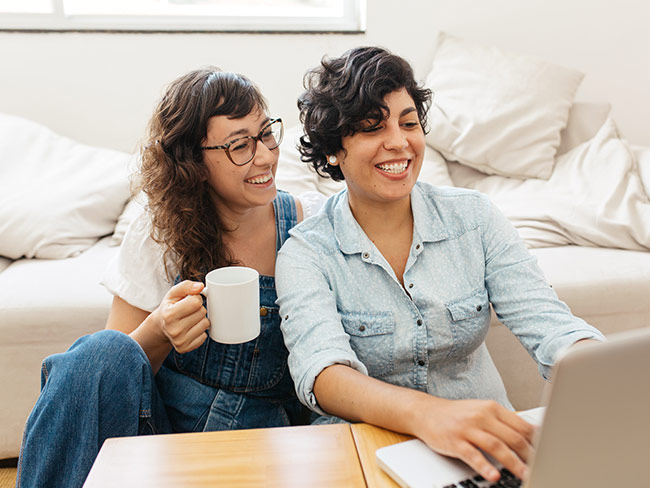Happy lesbian couple having coffee and using laptop in living room.