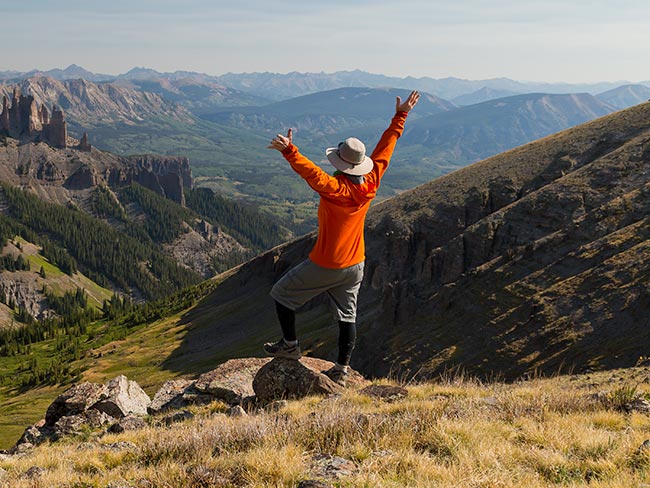 A man holding his arms in the air while he hikes on a ridge of a mountain with mountain peaks in the background