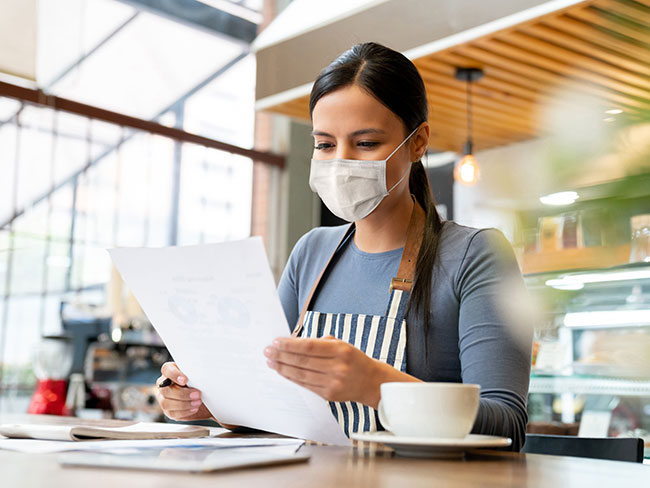 A Latina businesswoman wearing a face mask, in a coffee shop reading a document.