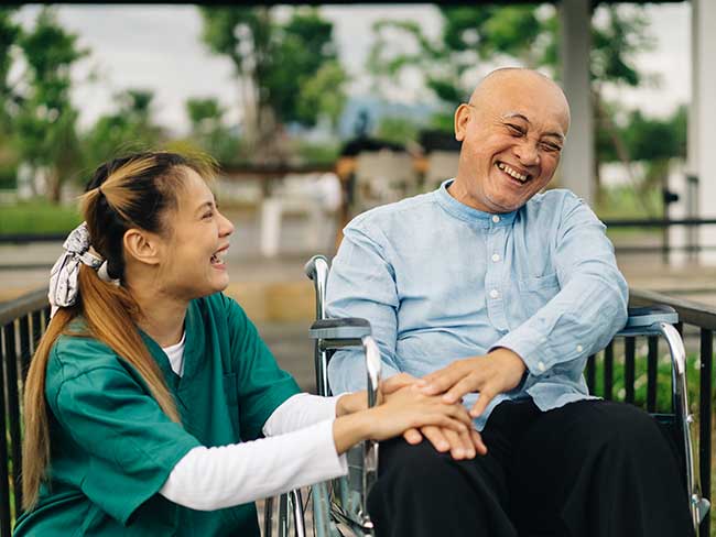 Nurse kneeling down next to a senior man in a wheelchair. They are laughing together. 