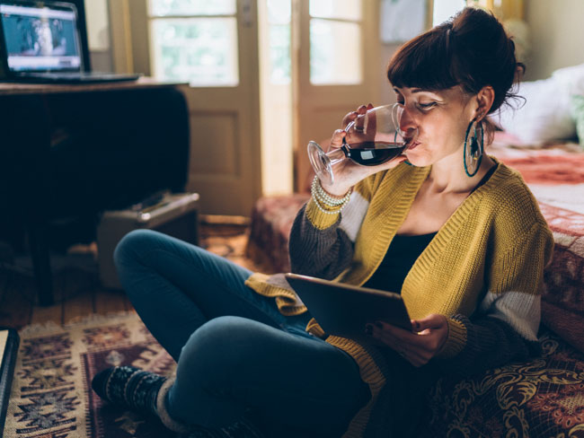 Woman casually drinking a glass wine at home while looking at her tablet computer.