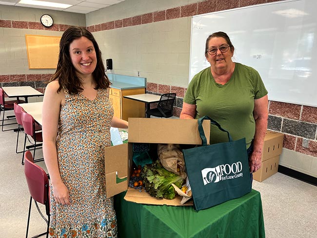 Emily Garver and Lisa Gray are standing next to a table with a box of fresh vegetables on it.