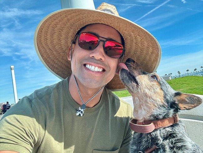 A man wearing a hat and sunglasses takes a selfie with his dog, who licks his face. They are at the beach.