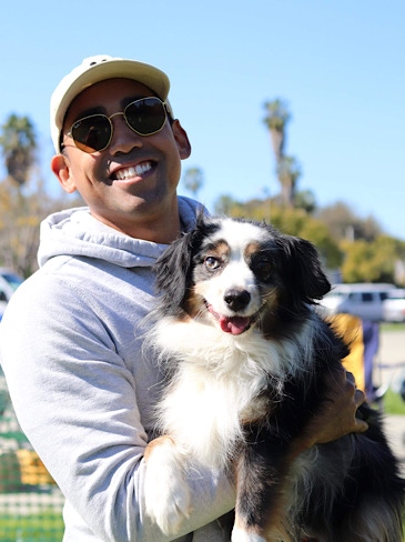A man wearing a hat and sunglasses holds up his dog for a photo while at the park.