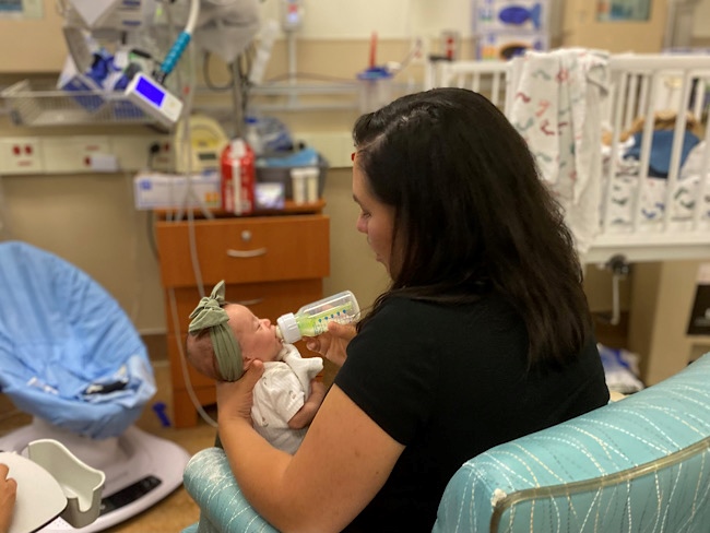 A woman in a hospital room sits in a chair and bottle feeds a tiny baby.