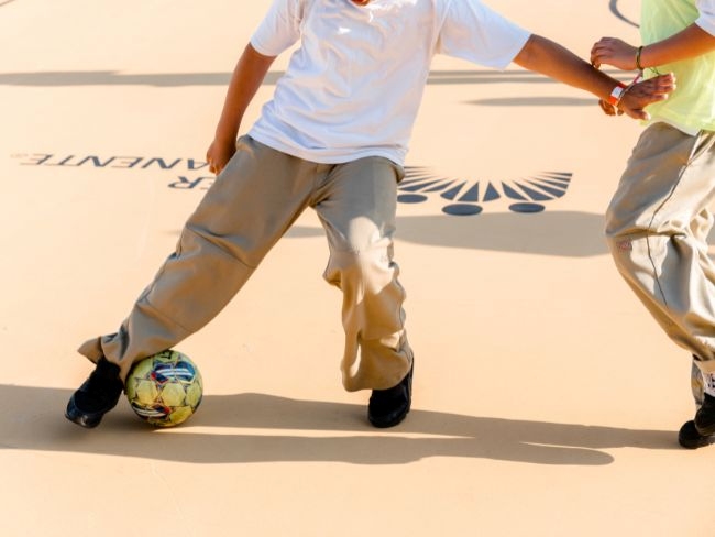Two children in khaki pants are playing soccer on an outdoor futsal court, with one child controlling the ball with their left foot.