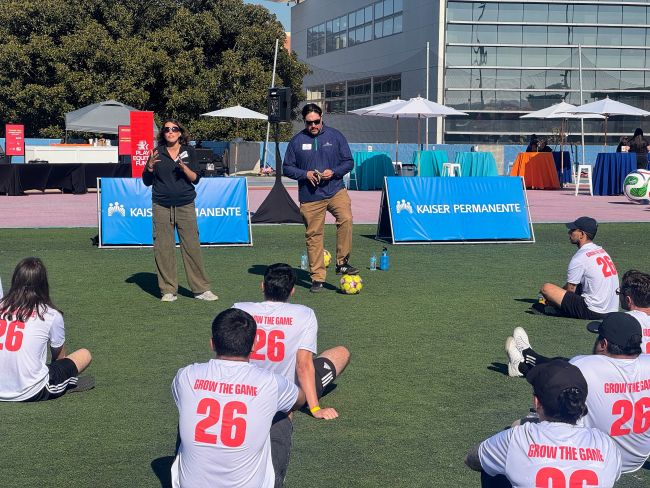 Two presenters, one standing with a soccer ball, address a seated group of participants wearing matching "Grow the Game 26' shirts on a turf field featuring Kaiser Permanente signage.