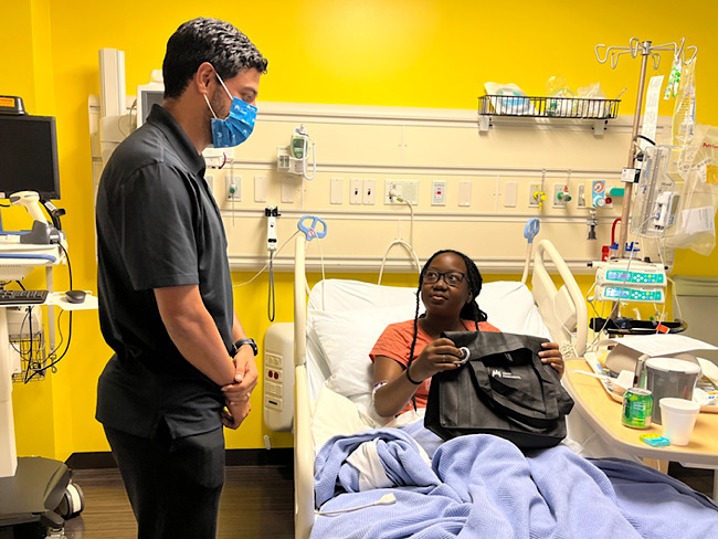 Carlos Vela, who is wearing a mask, stands next to the hospital bed of Lauren Lewis, a young girl. She is holding a bag given to her by Vela.