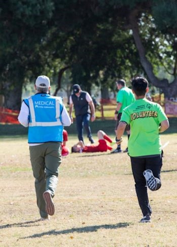 A person wearing a vest with the word "medical" on the back and another person wearing a t-shirt with the word "volunteer" on the back race toward a Special Olympics athlete who has fallen during a soccer match.