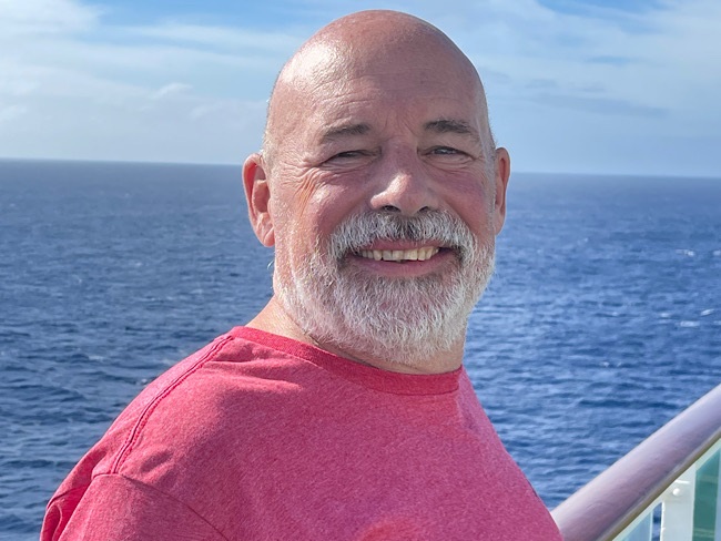 A man smiles for a picture. Behind him, the ocean stretches as far as the eye can see into a cloudy blue horizon.