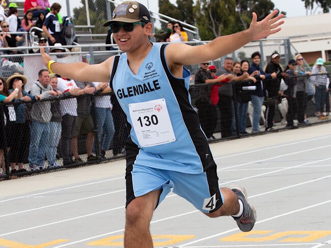 A young person runs on a track in front of a crowd while holding his arms out.