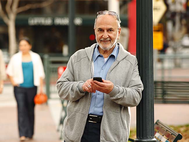 A senior man leaning against a pole, holding a mobile phone.