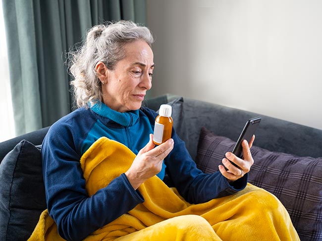 Mature female patient, under a blanket at home, shows her doctor the prescription medication she needs in an online call with her doctor using her mobile phone.