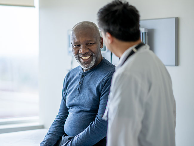 A smiling, older Black man talks with a doctor in a medical exam room.
