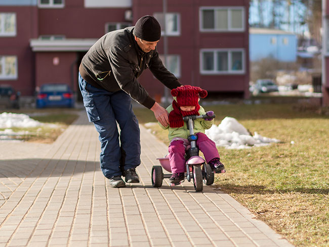 Grandfather teaching his toddler granddaughter how to ride a tricyle on a cold and sunny winter day.