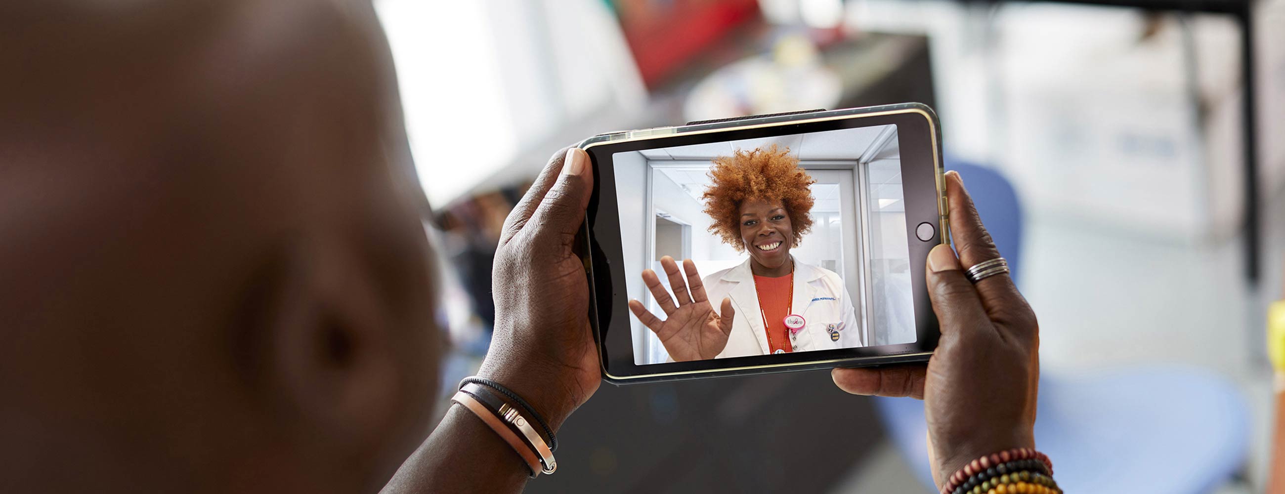 A smiling female physician waving while her patient holds a tablet during a telehealth visit.