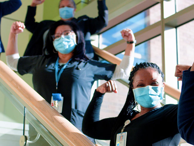 A group of people in blue, in a sunny stairwell wearing facemasks and flexing their arms. 