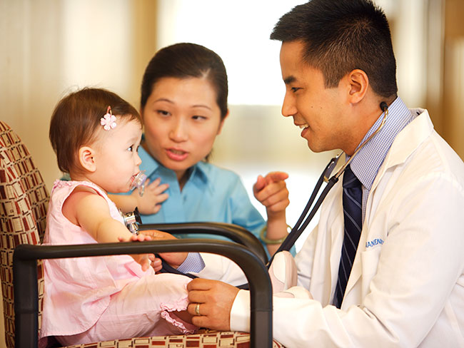 Male Asian American Kaiser Permanente physican talking to a cute baby girl dressed in pink while her mother gestures in the background. 