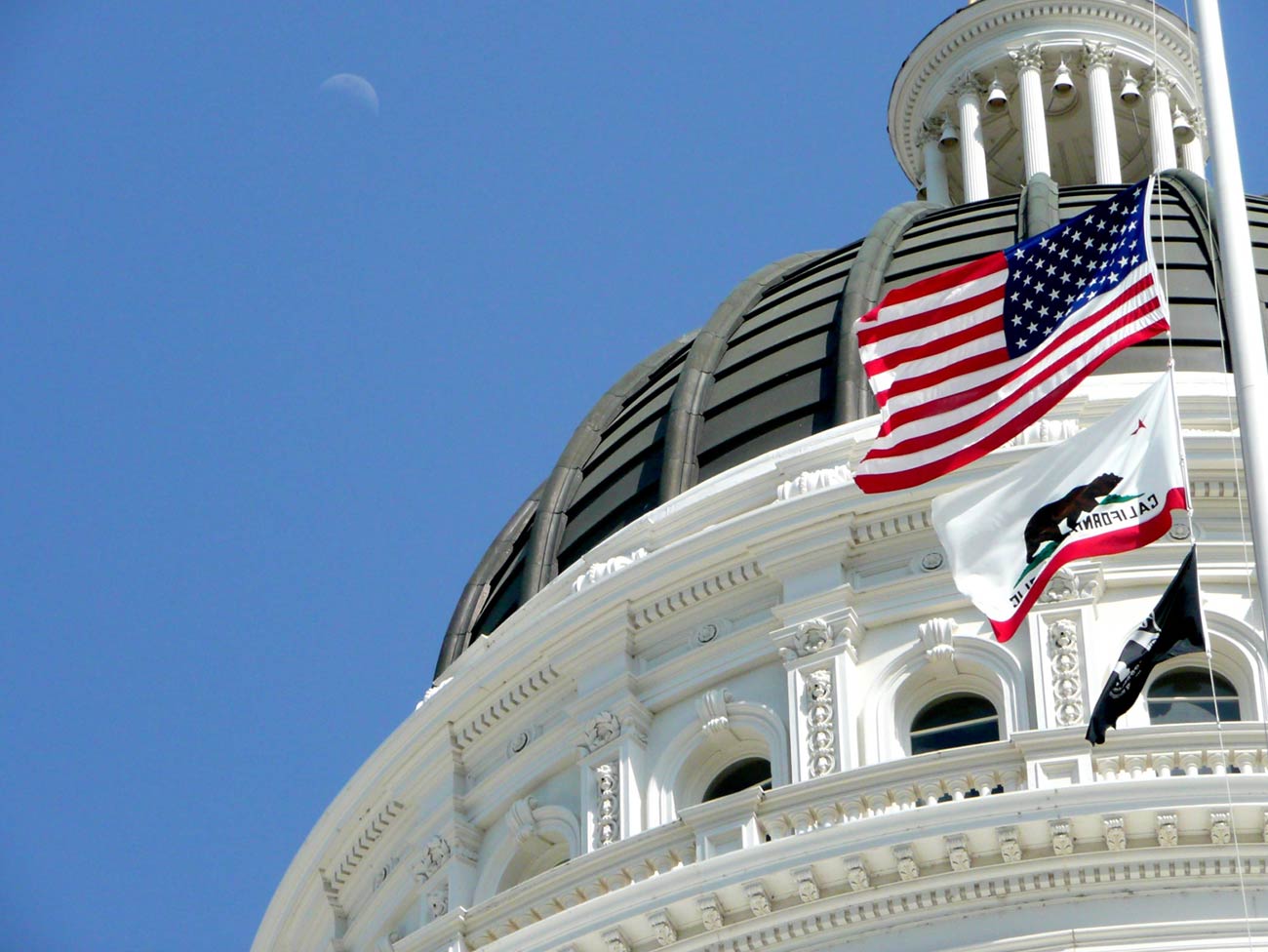 California Capitol building dome with the U.S. and California state flags flying in the wind in front of it.