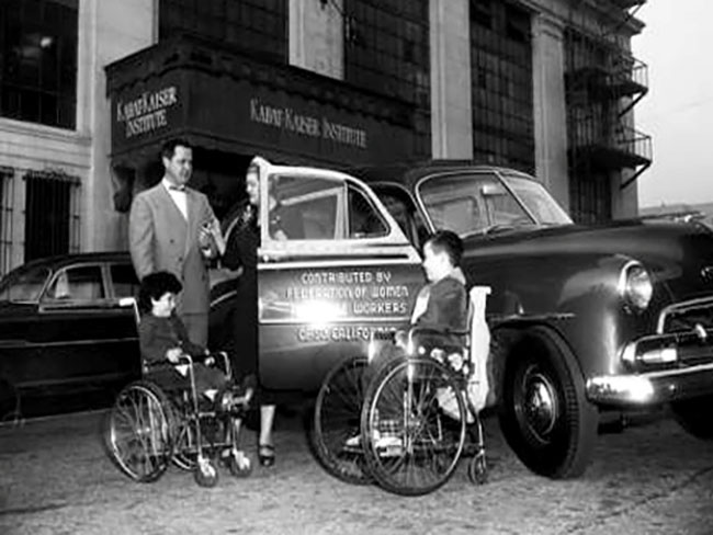 1952 image of a station wagon with a man and a woman standing next to the car along with two children in wheelchairs.