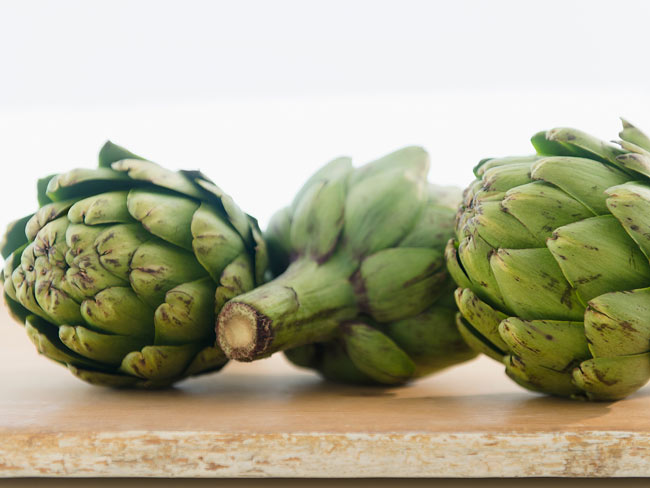 Three whole artichokes on top of wood table.